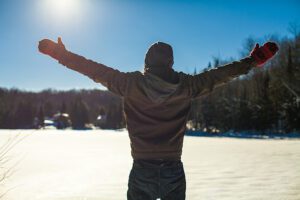 Person with winter coat, mittens and hat faced away toward snowy lake and forest